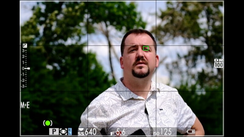 A man in a white patterned shirt stands outdoors with trees in the background, viewed through a camera viewfinder display. The camera settings are visible, and a green focus box highlights his left eye.