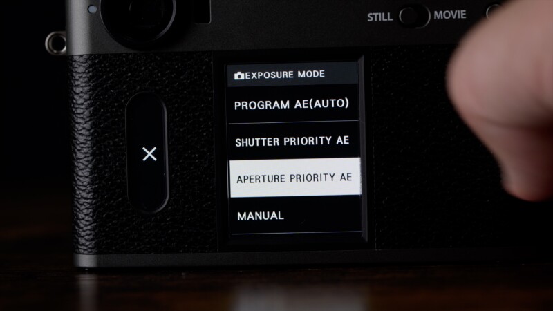 Close-up of a digital camera’s screen displaying exposure mode options: Program AE (Auto), Shutter Priority AE, Aperture Priority AE (highlighted), and Manual. A finger is selecting an option.