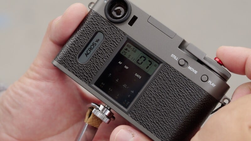 A close-up of a person's hand holding a retro-style digital camera with a textured grip, a display screen showing camera settings, and several buttons including “STILL,” “MOVIE,” and “PLAY.”.