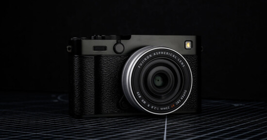 A black Fujifilm digital camera with a textured grip and aspherical lens is placed on a grid-patterned surface against a dark background.