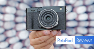 A hand holds a black camera in front of a blurred patterned background. The image includes the "Petapixel Reviews" logo in the bottom right corner.