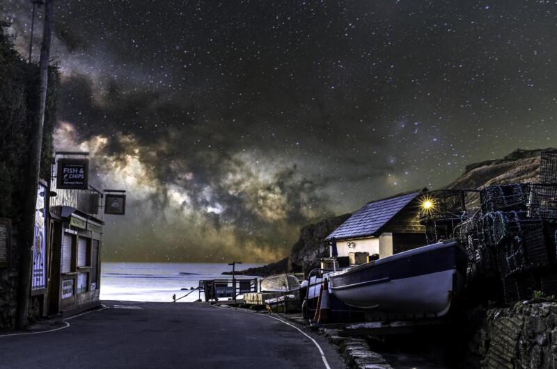 A narrow coastal road curves past a small boat shed and fishing nets, with boats on the side. The night sky above sparkles with stars and the bright arc of the Milky Way, creating a dramatic contrast over the calm sea.