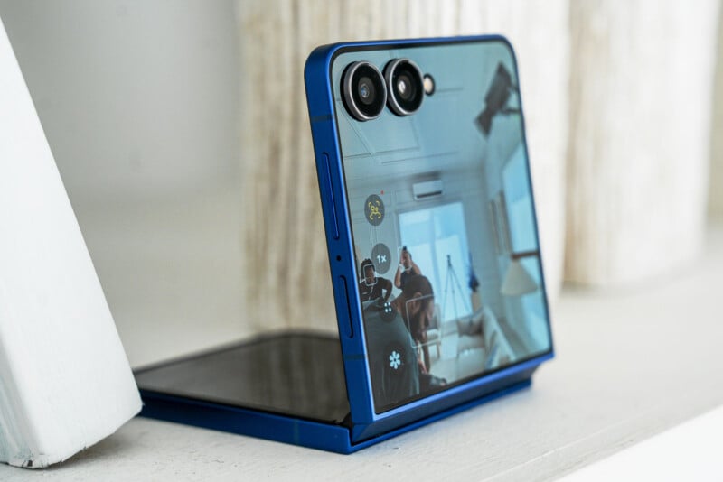 A blue foldable smartphone is partially open on a white surface, with its external screen displaying the camera app and reflecting a person sitting in a bright, modern room.