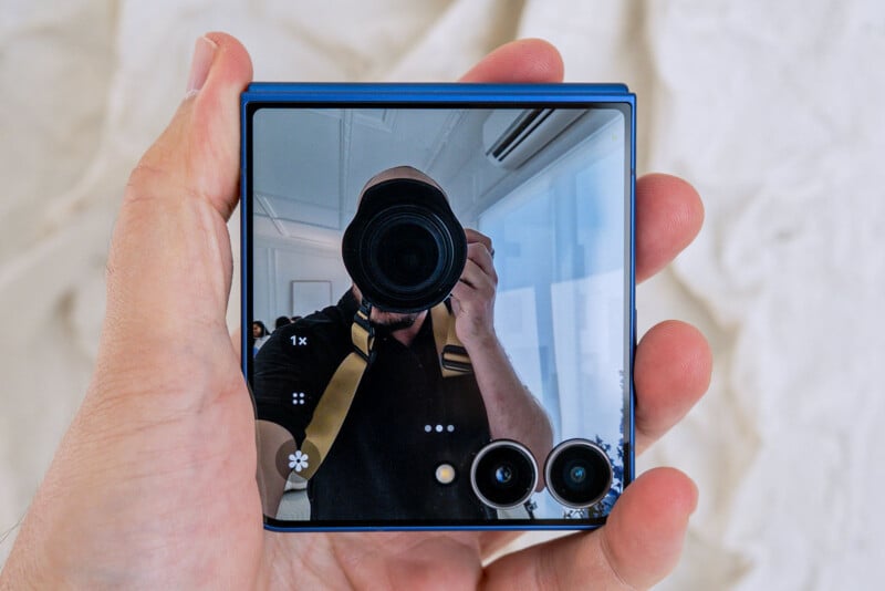 A person holding a folded smartphone with a reflective screen, showing their reflection as they take a photo. The phone has two camera lenses and is blue in color. The background is blurred.