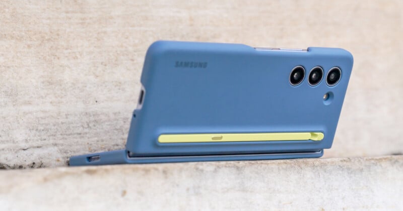 A blue Samsung phone with a yellow accent in its case is propped up horizontally on a light stone surface. The back shows three camera lenses and a small round flash.