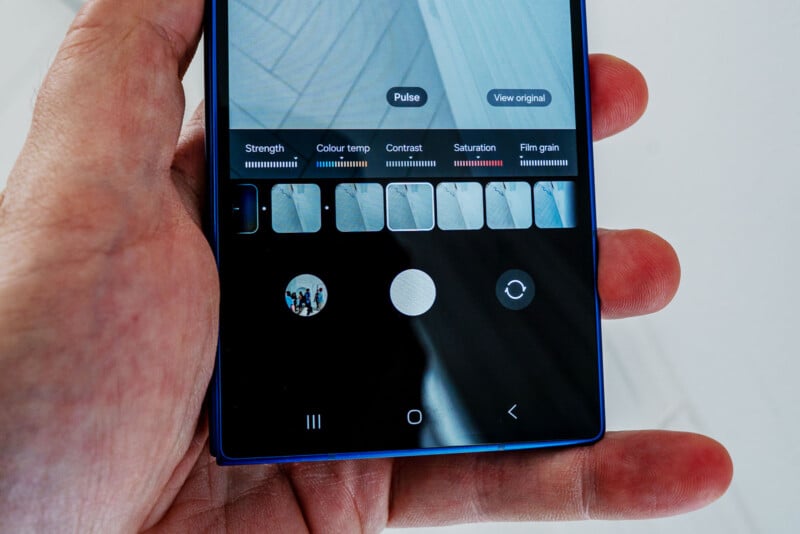 A hand holds a smartphone displaying a photo editing screen with filter options, adjustment sliders, and icons for strength, color temperature, contrast, saturation, and film grain.