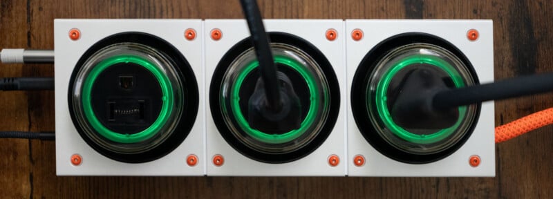 A close-up of three white power outlets with black plugs inserted, each surrounded by a glowing green ring, mounted on a wooden surface. Orange screws secure the outlets in place.