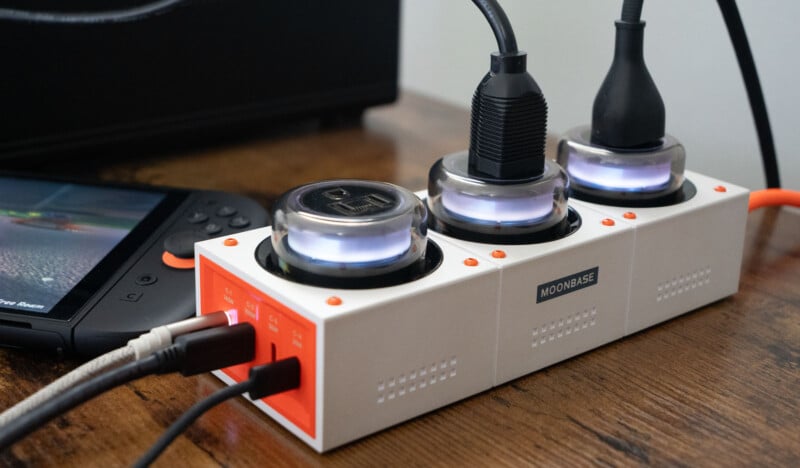 A white and orange power strip labeled "MOONBASE" on a wooden surface, with three round plugs connected and glowing, next to an electronic device with a screen and controller on the left.