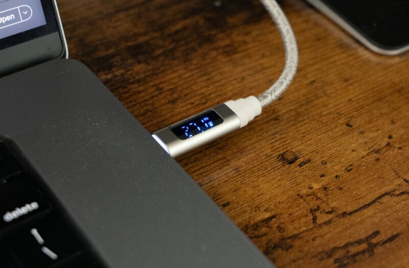 A braided charging cable with a small digital display showing "21W" is plugged into a laptop on a wooden surface.