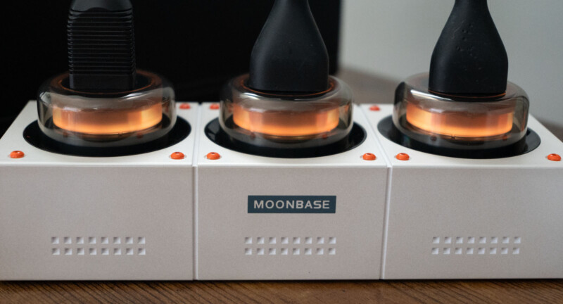 Three black vape devices sit in illuminated charging docks labeled "MOONBASE," with a wooden surface beneath.