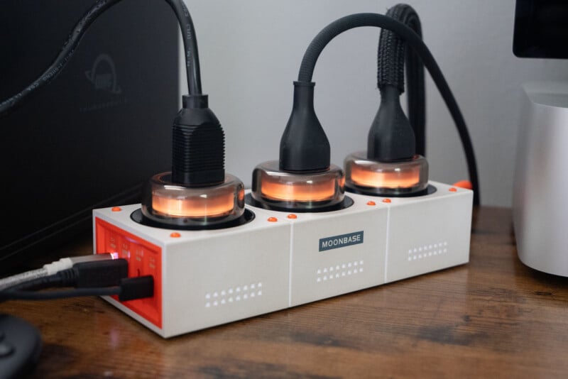 A white and orange power strip labeled "MOONBASE" with three large black plugs connected. The power strip sits on a wooden desk next to computer equipment.