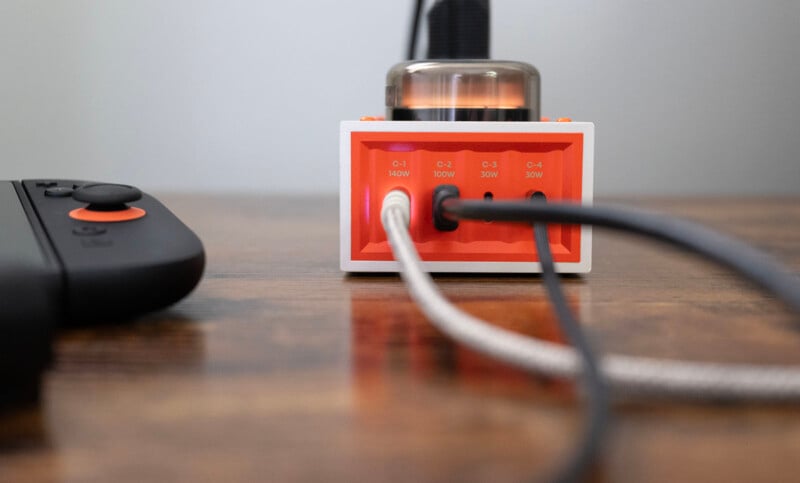 A close-up of a small orange and white charging hub with multiple cables plugged in, sitting on a wooden surface. A game controller is partially visible on the left side of the image.