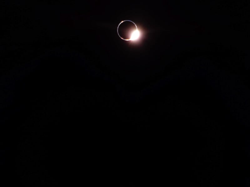 A solar eclipse with a bright ring of sunlight peeking from behind the moon against a dark sky, creating a "diamond ring" effect. The light appears as a small burst on one side of the moon's silhouette.