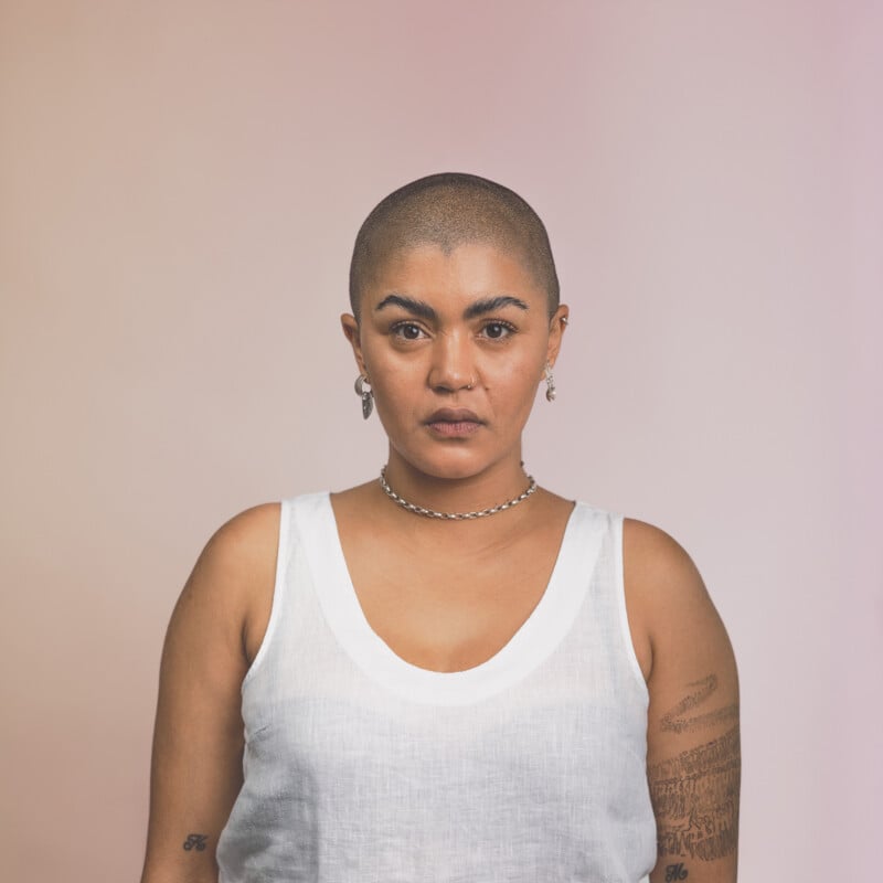 A person with a shaved head, wearing a white sleeveless top and silver jewelry, stands against a gradient pink and beige background, looking directly at the camera. Tattoos are visible on their right arm.