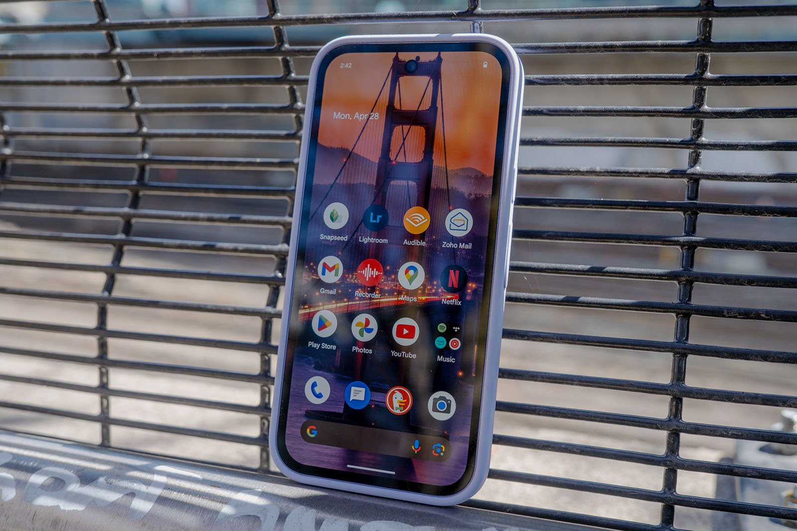 A smartphone leaning against a metal bench displays its home screen with various app icons; the screen shows a wallpaper of the Golden Gate Bridge.
