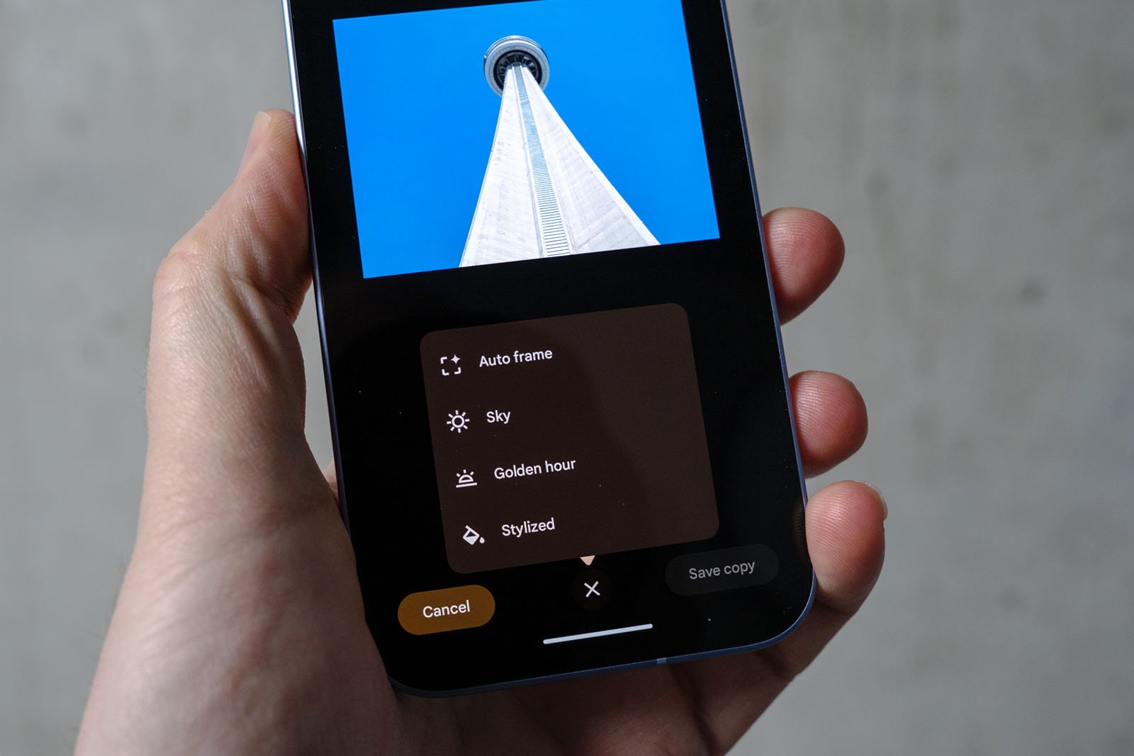 A hand holds a smartphone displaying a photo editing app. The screen shows a photo of a tall tower against a blue sky and an editing menu with options like auto frame, sky, golden hour, and stylized.