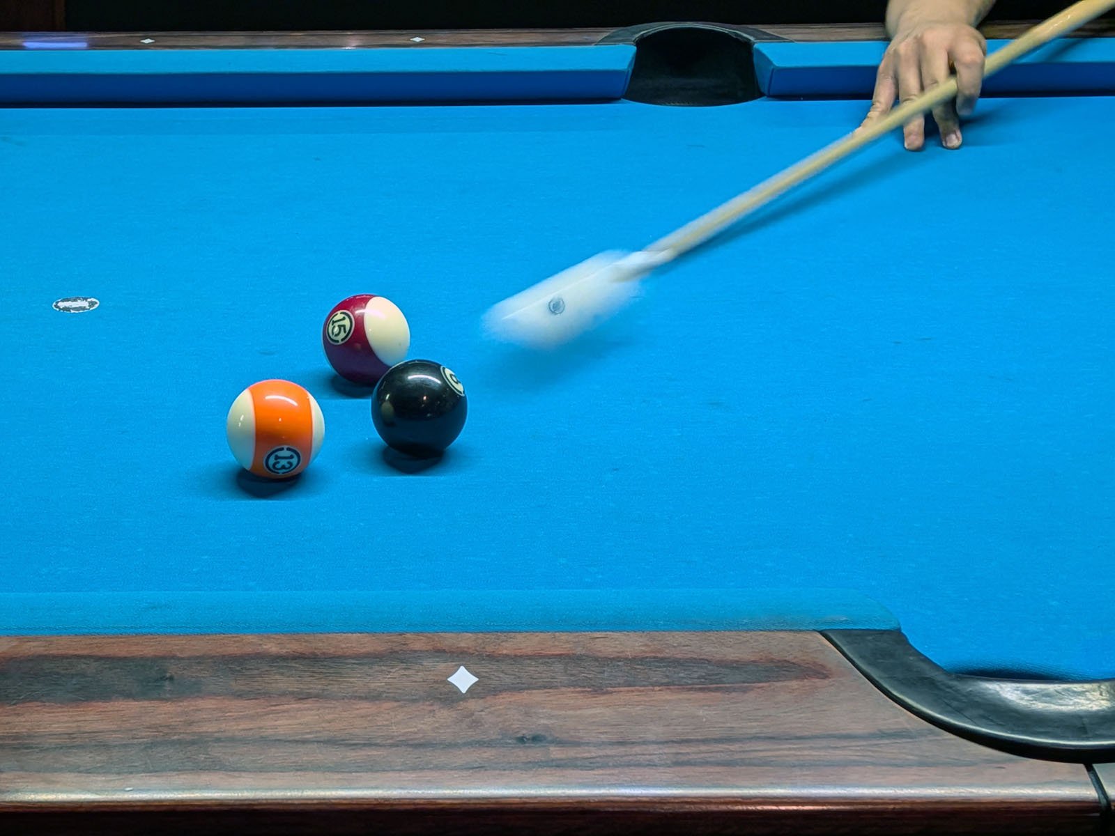 A person’s hand is holding a cue stick, striking the cue ball on a blue pool table. Three other balls, numbered 9 (yellow and white), 15 (red and white), and 8 (black), are grouped together nearby.