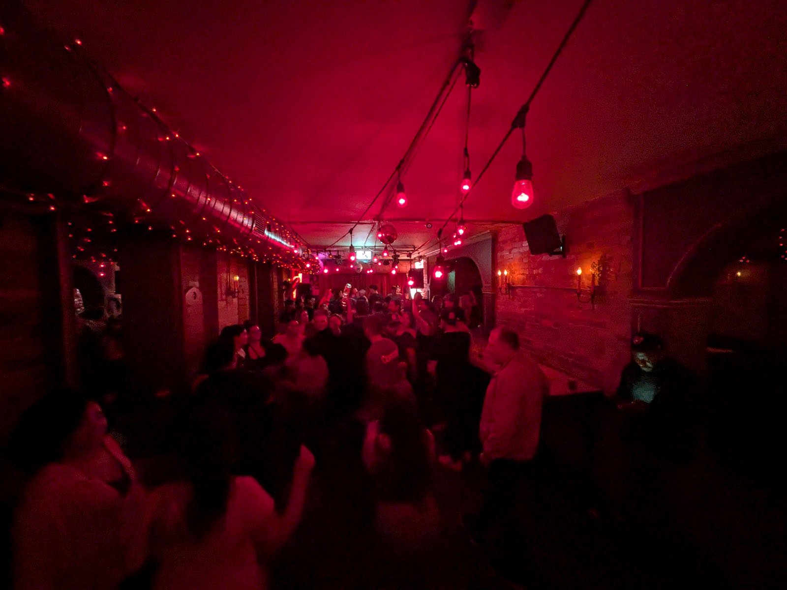 A dimly lit bar or club with red lighting, crowded with people dancing and socializing. Strings of red lights hang from the ceiling, and the atmosphere appears lively and festive.