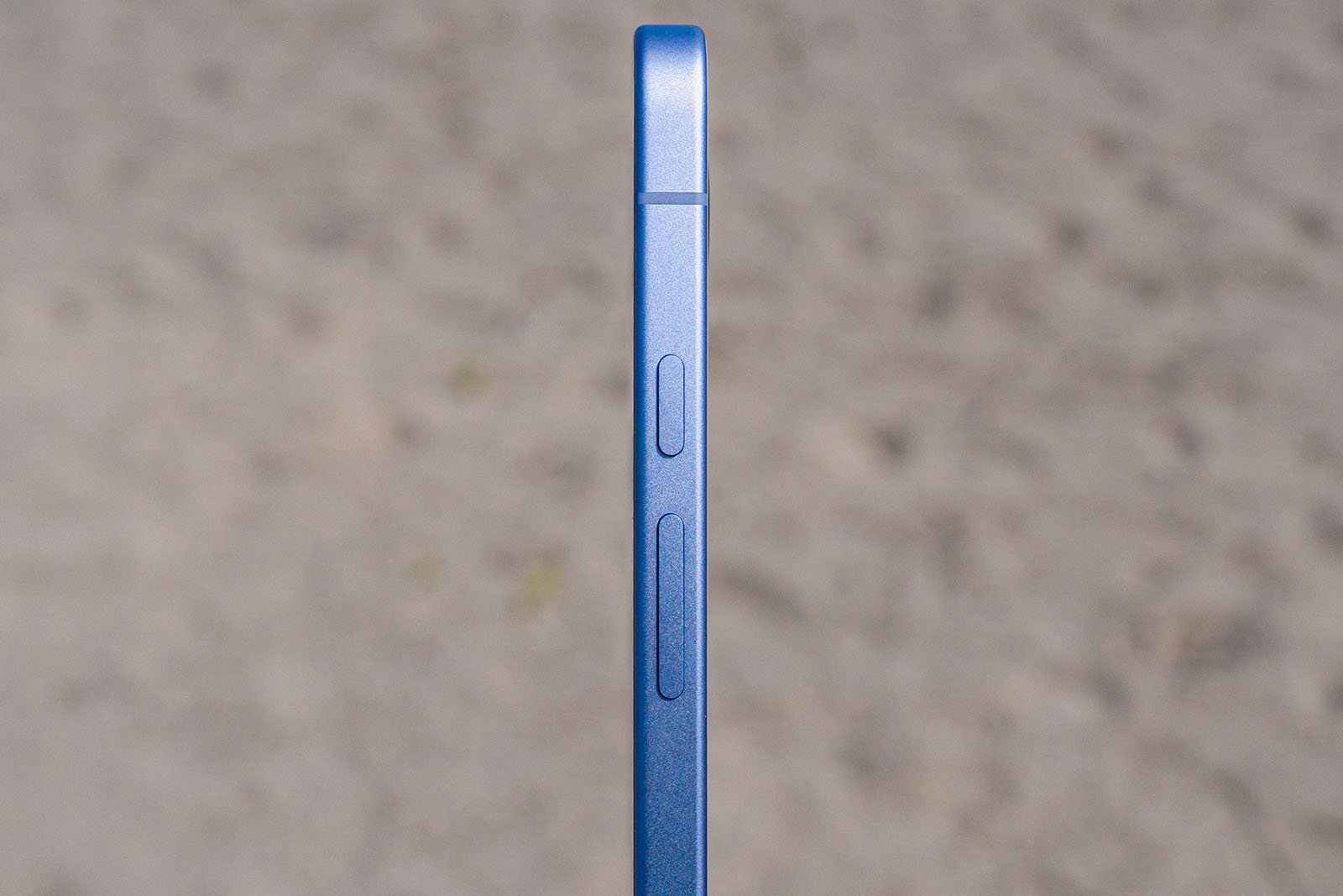 A close-up side view of a blue smartphone showing its volume buttons and a switch, with a blurred neutral background.