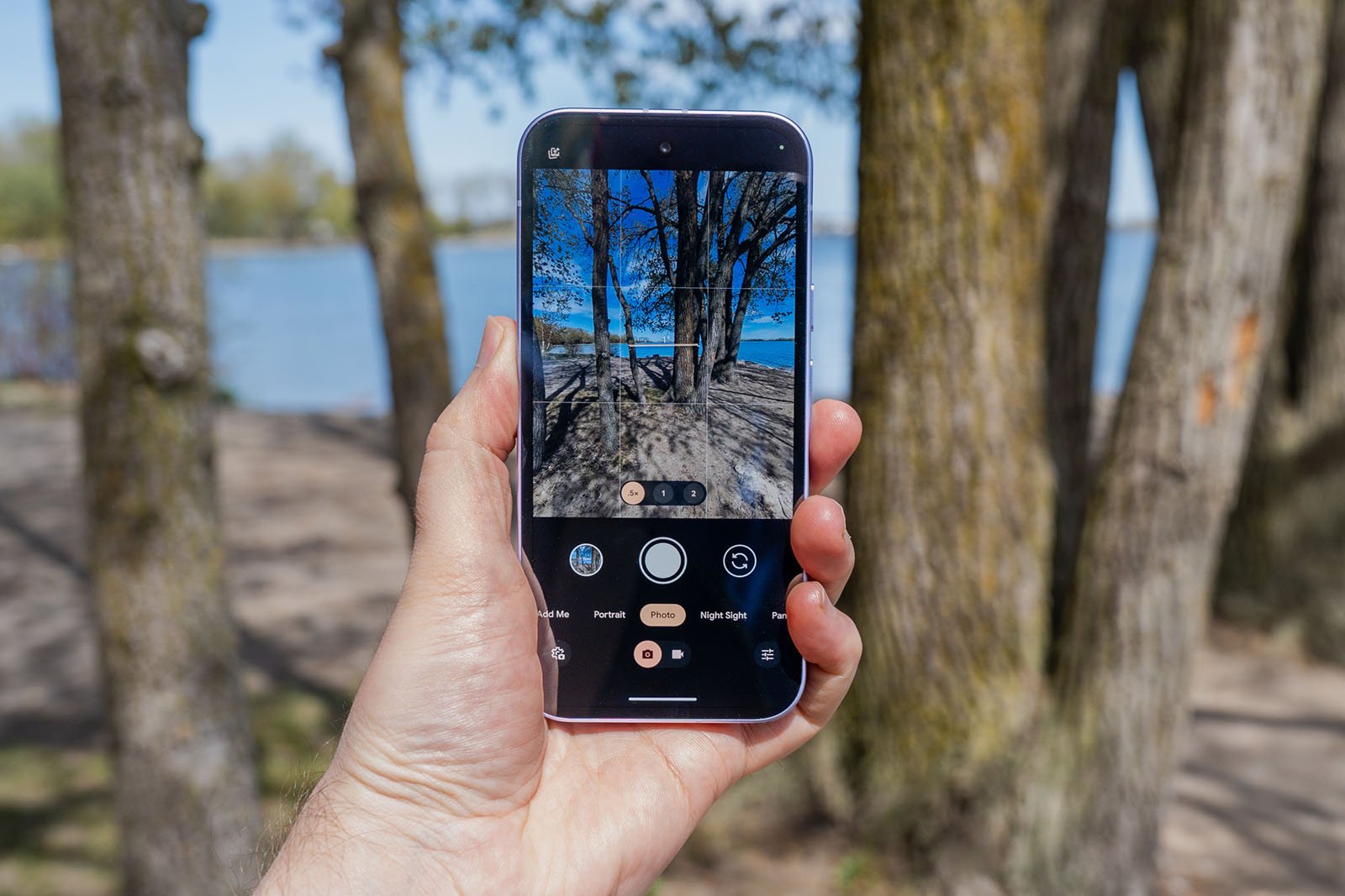 A hand holds a smartphone, taking a photo of trees and a lake in a park. The phone screen displays the camera app with the same scene in view, showing clear blue sky and water in the background.