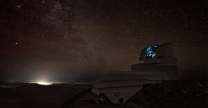 A large astronomical observatory sits atop a mountain under a star-filled night sky. In the distance, city lights glow, while the observatory’s dome is partially open, illuminated with a faint blue light inside.