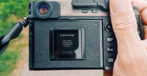A close-up of a person holding a digital camera, showing its screen displaying a “Product Planning” credits list with four names. The camera’s buttons and textured grip are visible, with a blurred outdoor background.