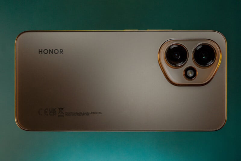 A gold-colored Honor smartphone is shown from the back, featuring dual cameras and a flash in a rounded rectangular camera module, with branding and certification symbols visible.