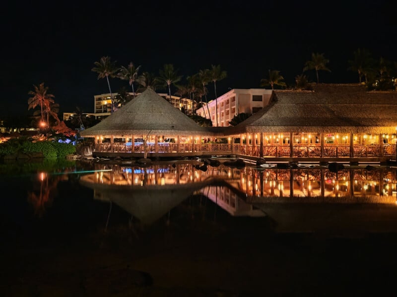 Thatched-roof buildings and palm trees are illuminated at night, reflecting on a calm body of water, with hotel structures visible in the background.