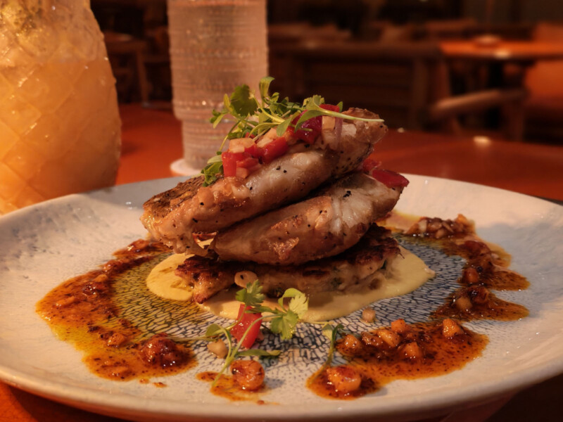 A dish featuring two grilled fish fillets stacked on top of vegetable cakes, garnished with microgreens and diced red peppers, served with a creamy yellow sauce and spicy oil on a white plate.