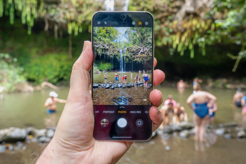 A hand holds a smartphone, capturing a photo of people standing and swimming near a waterfall in a lush, green outdoor setting. The phone screen shows the scene being photographed.