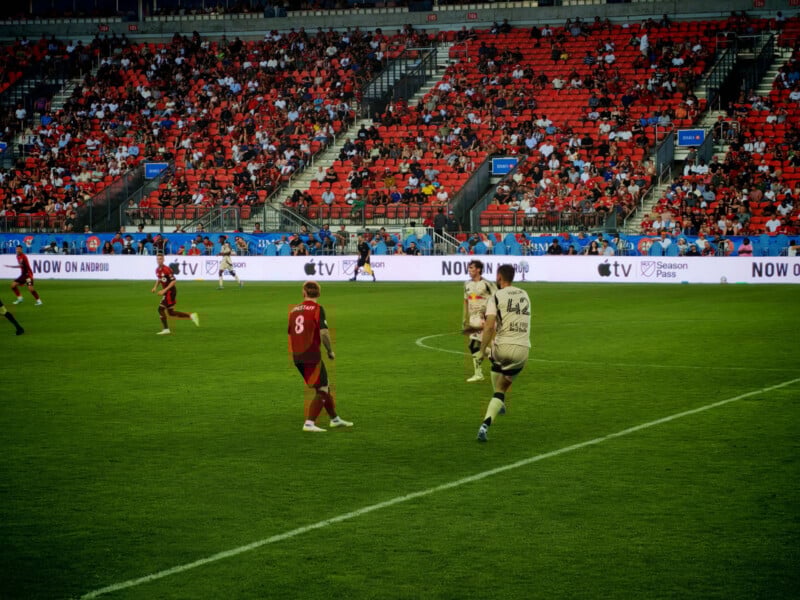 Soccer players in red and beige uniforms play on a green field, with a packed stadium of spectators and digital ads visible in the background.