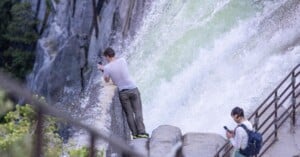 A man leans on a railing close to a powerful waterfall, taking photos, while a woman with a backpack stands on a nearby path looking at her phone. Water splashes against rocks, and greenery surrounds the scene.
