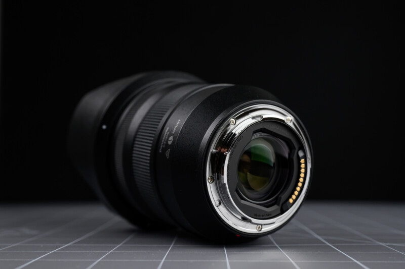 A close-up of a camera lens lying on its side on a gray tiled surface, showing the metal lens mount and electronic contacts, with a dark, blurred background.