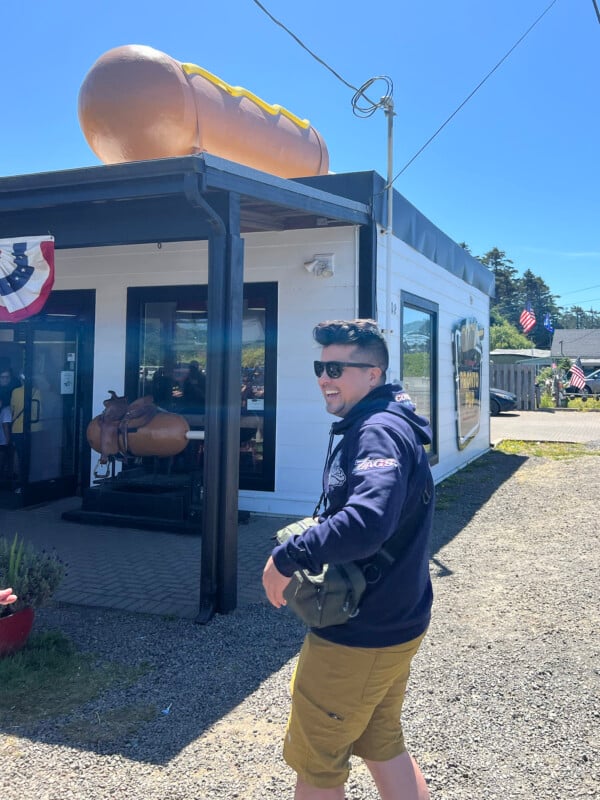 A man wearing sunglasses, a dark hoodie, and yellow shorts smiles while walking outside a small hot dog restaurant with a large hot dog sculpture on its roof. It’s a sunny day with clear blue skies.