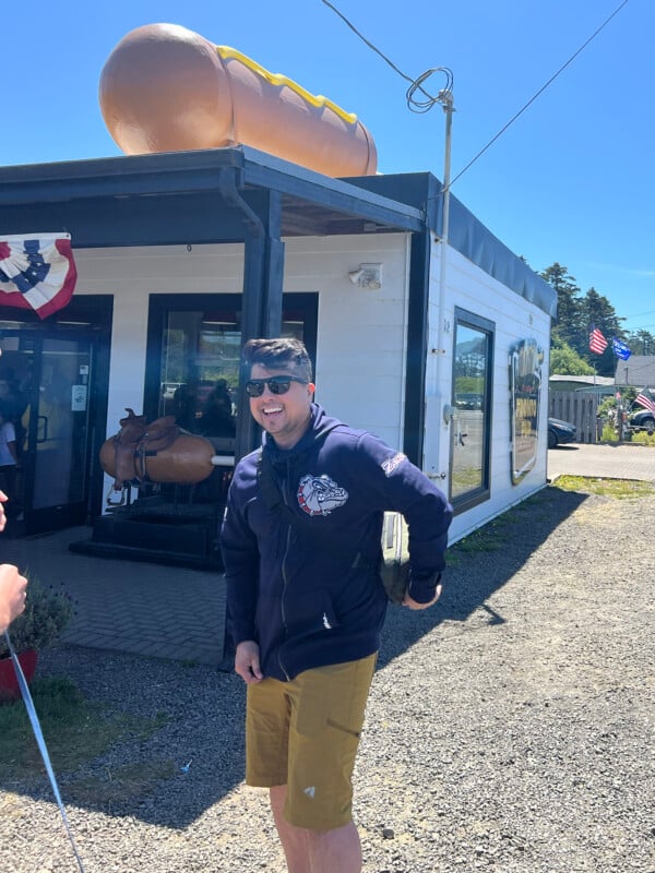 A man in sunglasses and a dark jacket stands smiling outside a white building with a large hot dog sculpture on the roof. The day is sunny, and American flags and festive decorations are visible.