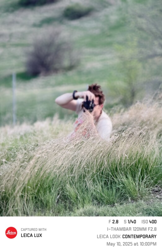 A person crouches in tall grass, holding a camera up to their face as they take a photo outdoors. The background is green and blurred, with camera settings and date info overlaid at the bottom.
