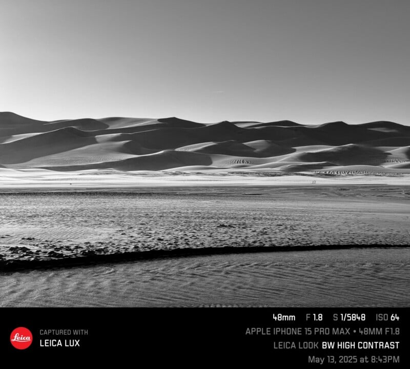 Black-and-white photo of vast sand dunes under a clear sky, with smooth ridges and scattered shadows. In the foreground, textured sand appears darker, creating strong contrast. Image info overlay is visible at the bottom.