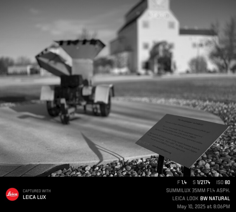 A black and white photo of a small, wheeled artillery piece outdoors, with an informational plaque in the foreground and a tall building out of focus in the background. Camera details are displayed at the bottom.