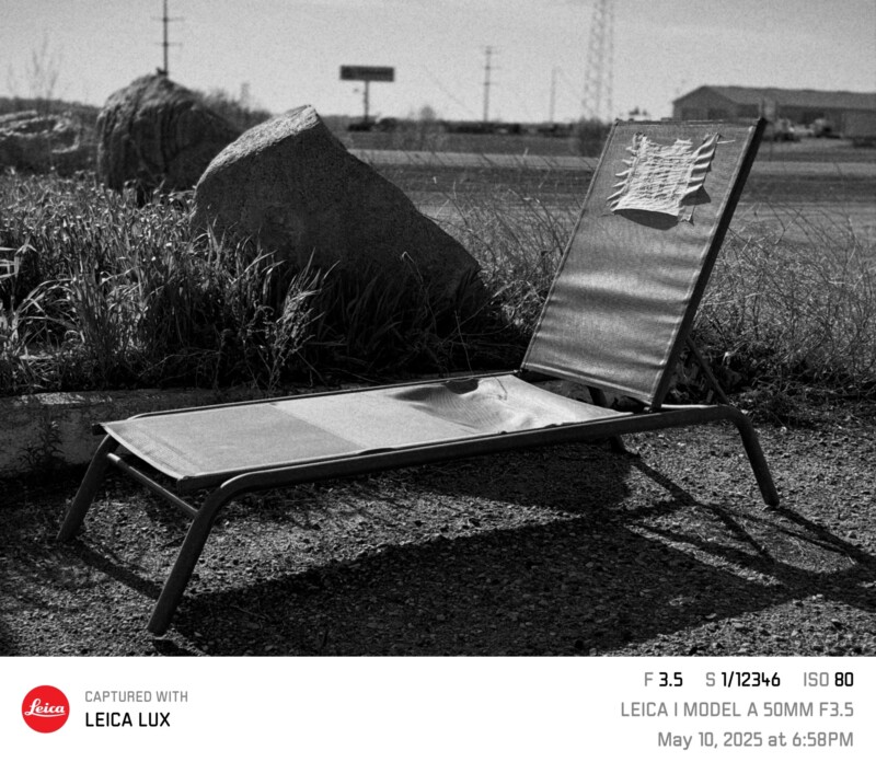 A lone outdoor lounge chair sits beside tall grass and a large rock on a paved surface under cloudy skies. Shadows are long, and a paper is attached to the chair's back. The photo includes camera settings and a Leica logo.