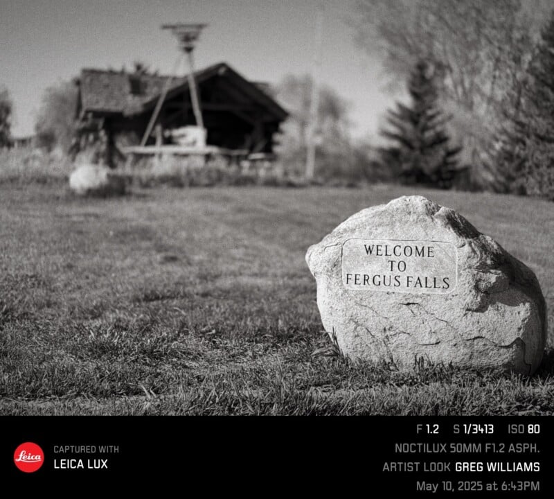 A stone with the inscription "Welcome to Fergus Falls" sits in a grassy area. In the blurred background, there is a wooden structure with a small roof and trees. The image details and photographer info appear at the bottom.