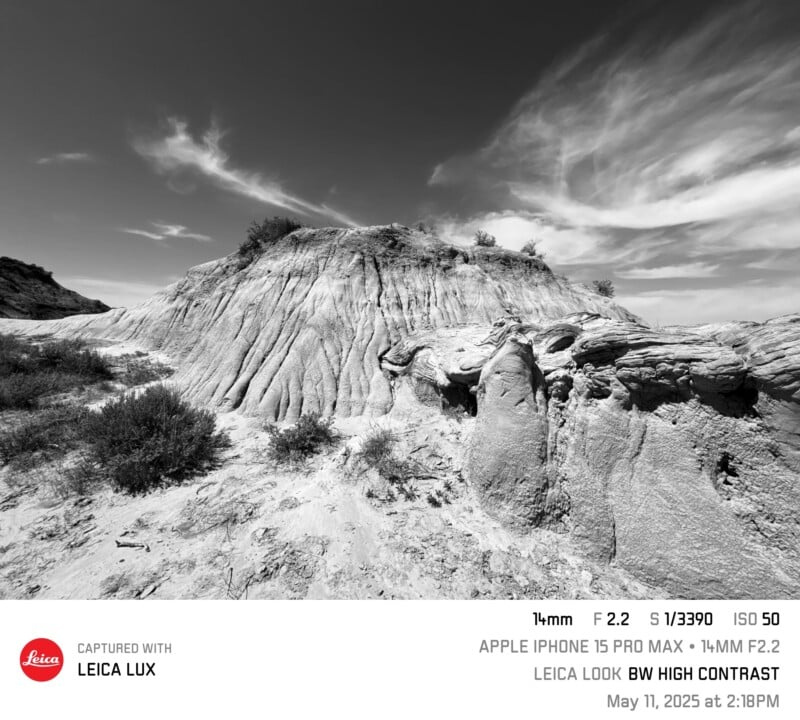 Black and white photo of dramatic, eroded rock formations under a sky with wispy clouds. Sparse vegetation grows atop the rocks and on the ground. The scene appears rugged and desolate.