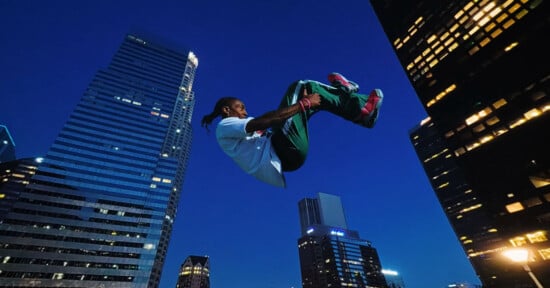 A person in mid-air performing a backflip at night, surrounded by tall city skyscrapers with illuminated windows against a dark blue sky.