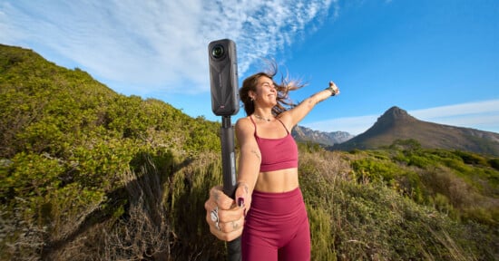 A woman in a maroon workout outfit smiles and holds a 360-degree camera on a selfie stick in a scenic outdoor setting with green hills and a mountain under a blue sky with wispy clouds.