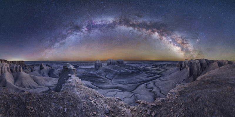 A stunning nightscape of a rugged, eroded canyon under a star-filled sky, with the Milky Way forming a bright, colorful arc above the rocky landscape.
