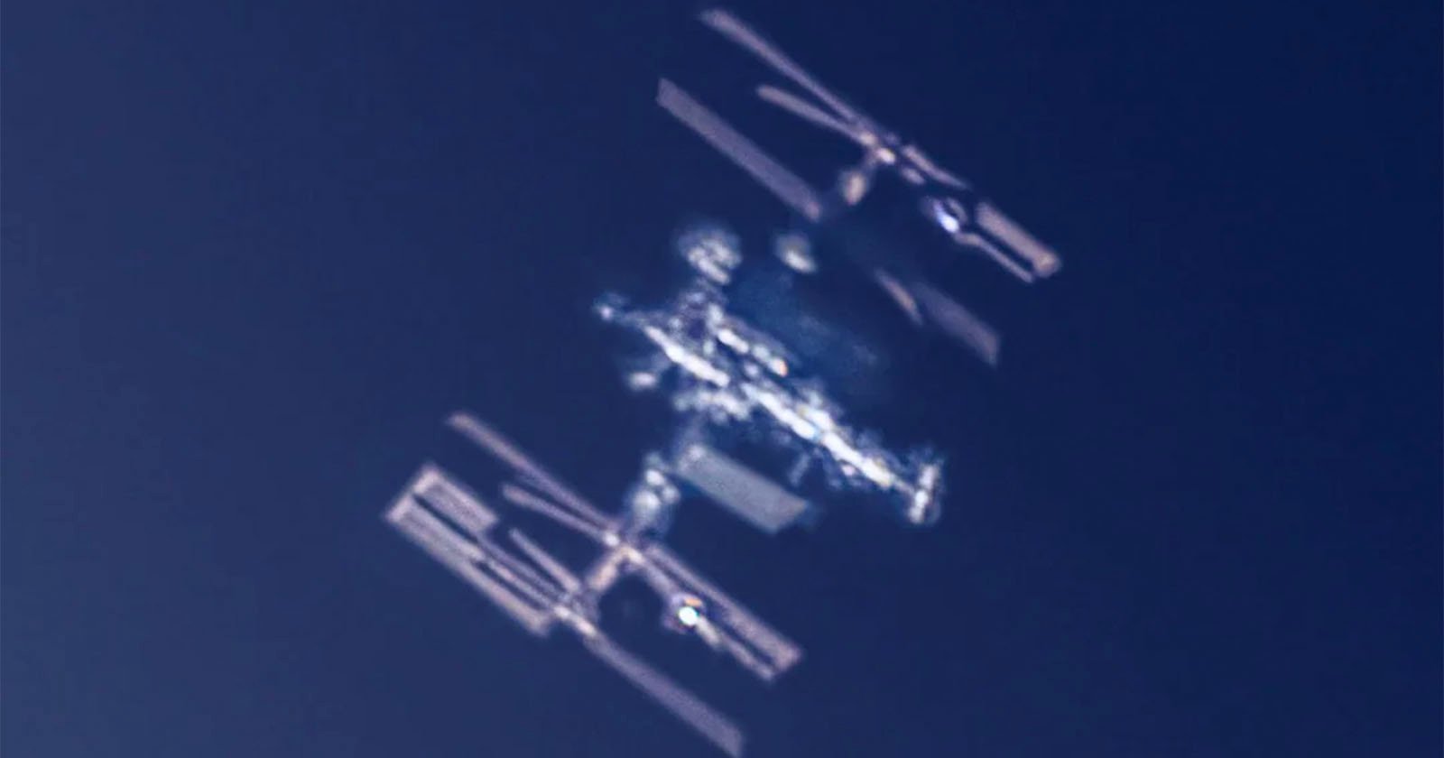 A blurred, close-up image of the International Space Station (ISS) orbiting against a dark blue sky, with its solar panels and structural modules visible.