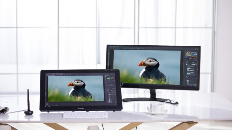 A desk with a digital drawing tablet and a monitor displaying the same image of a puffin. A stylus, keyboard, mouse, notebook, and a white coffee cup are also on the desk, with large windows in the background.
