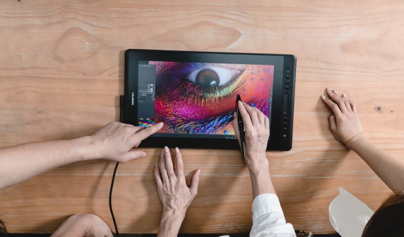 Three people work on a digital drawing tablet displaying a colorful close-up of an eye. One uses a stylus to edit the image, while the others use their hands to interact and assist with the design process.