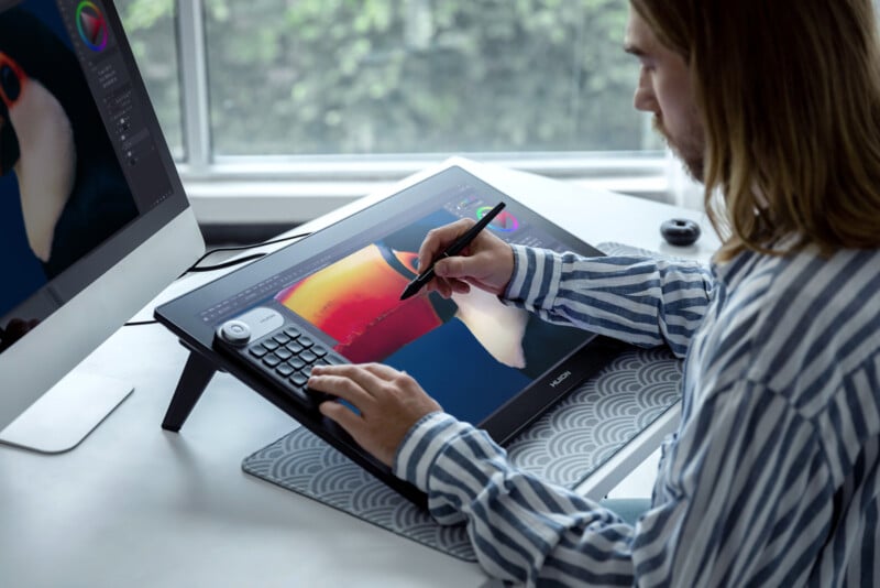 A person with long hair uses a digital pen and drawing tablet to create digital art, with a colorful artwork displayed on the screen. A desktop computer and window with greenery are visible in the background.