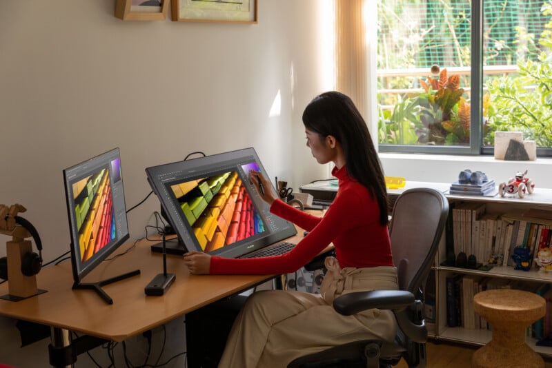 A woman in a red top works on a digital drawing tablet at a desk with two monitors in a bright home office, with bookshelves, artwork, and a window overlooking a garden.