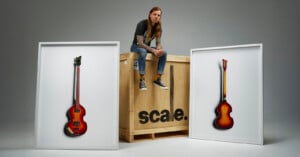 A man with long hair and tattoos sits on a large wooden crate labeled "scale." Two framed photos of vintage guitars are displayed on either side of the crate against a plain gray background.